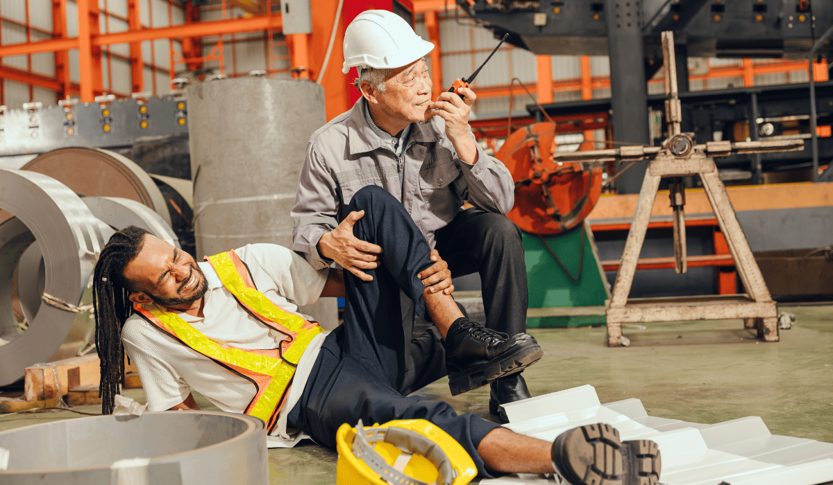 Industrial worker injured on the factory floor being assisted by a colleague who calls for help, illustrating a workplace lifting injury in Schuylkill County, PA